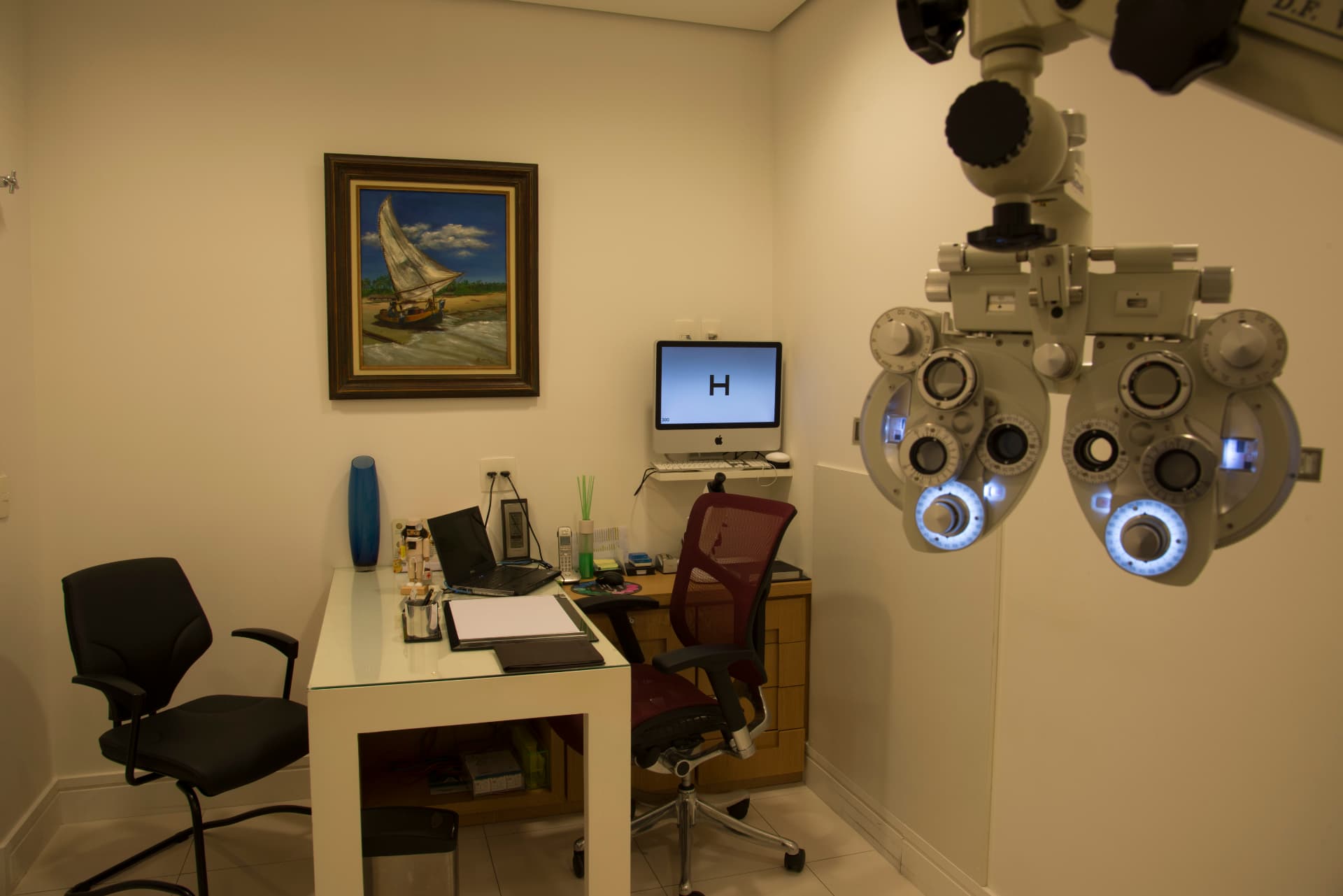 Consultório com refrator, mesa de atendimento e tabela de acuidade visual